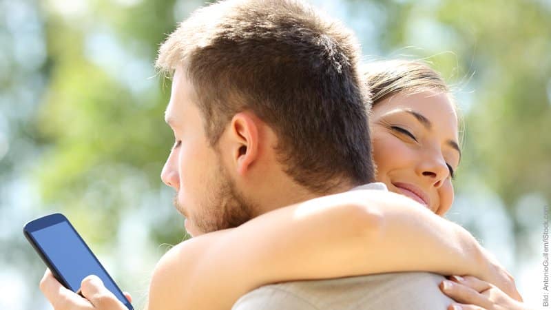Gefahr für die Beziehung durch das Internet. Mann und Frau umarmen sich. Er schaut hinter dem Rücken der Frau heimlich in sein Handy.