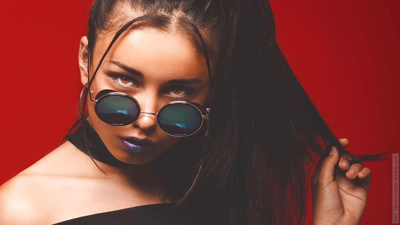 Warum lügen Partner sich an? Dunkelhaarige Frau schaut vielsagend über ihrer Sonnenbrille hindurch, zwirbelt sich dabei eine lange Haarsträhne. Sie glaubt wohl kein Wort von dem, was sie hört und geht davon aus, dass sie angelogen wird.